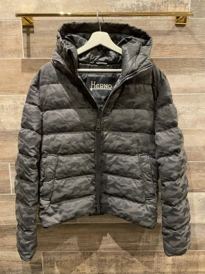 Herno jacka  - Snygg pufferjacka från Herno i grått camomönster. Jackan har huva, dragkedja framtill och quiltad design. Perfekt för kalla dagar med sin vadderade och isolerande känsla. Stilsäker och modern look som sticker ut. L passar M