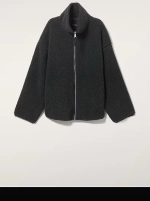 Svart teddyjacka med dragkedja - Mysig svart teddyjacka med hög krage och rak passform. Jackan har en stilren design med breda ärmar och dragkedja framtill. Tillverkad i fluffigt teddy-material som ger en trendig look.