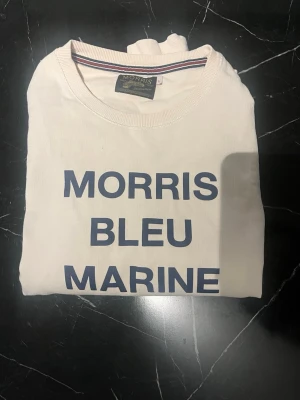 Vit sweatshirt från Morris - Vit sweatshirt från Morris med trycket 'MORRIS BLEU MARINE' i mörkblått framtill. Tröjan har rund halsringning, ribbade muddar vid ärmslut och nederkant samt en klassisk, avslappnad passform. Storlek L passar S-M. Har en liten fläck på bröstet 
