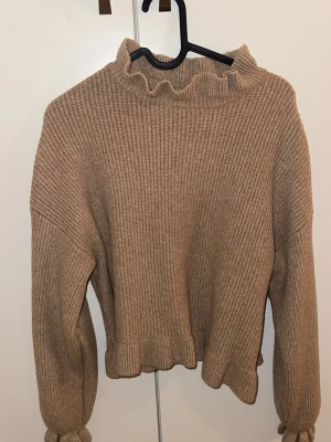 Beige stickad tröja från Primark - Mysig beige stickad tröja från Primark med ribbad struktur. Tröjan har volangdetaljer vid halsringningen och nederkanten samt långa ärmar med lätt puff och resår vid ärmslut. Perfekt för kyliga dagar och enkel att styla till olika outfits.