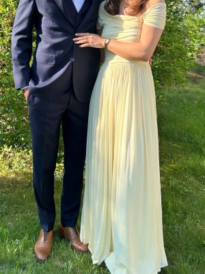 Ljusgul offshoulder långklänning - Elegant ljusgul långklänning med offshoulder-ärmar och draperad överdel. Kjolen faller mjukt och har ett luftigt, svepande tyg som ger en romantisk känsla. Perfekt för sommarens speciella tillfällen.