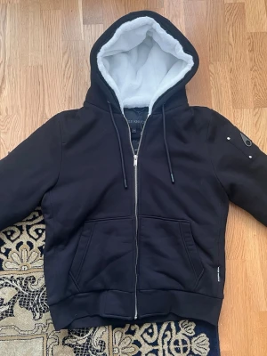 Moose Knuckles Bunny hoodie i storlek S - Helt Ny - Denna Moose Knuckles Bunny hoodie i storlek S är en perfekt balans mellan stil och funktion. Med en mjukt fodrad huva och en stilren zippered design håller den dig varm och bekväm. Hoodiejackan har praktiska fickor och moderna detaljer på ärmen, vilket ger en sportig och trendig look. En mångsidig jacka som passar både för avslappnade dagar och kyligare väder.