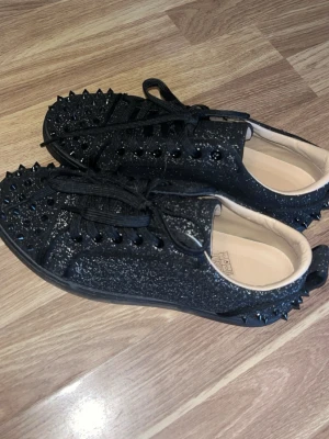 Svarta glittriga sneakers med nitar - Unika svarta sneakers från Christian Louboutin med glittrig finish och svarta nitar över hela skon. Lågt skaft, svarta skosnören och en chunky svart sula. Insidan är beige och materialet är en mix av läder och textil. Säljer då jag råka beställa i fel storlek.. Använda en gång och så gott som nya. 
