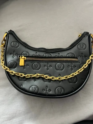 Svart axelväska från Vezzano  - Säljer en svart axelväska från Vezzano med präglad logga över hela väskan. Väskan har en snygg guldfärgad kedja som axelrem och dragkedja upptill samt en extra ficka med dragkedja på framsidan. Tillverkad i skinn och har gulddetaljer.