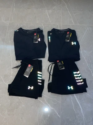 Svart träningsset från Under Armour - Snyggt svart träningsset från Under Armour med t-shirt och shorts. Setet har reflekterande loggor och detaljer i regnbågsfärger på bröst, ärm och ben. Materialet är lätt och andas, perfekt för träning eller sportaktiviteter.