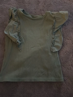 Grön ribbad topp med volangärm - Superfin grön ribbad topp från Kappahl med volangdetaljer längs axlarna. Toppen är ärmlös med breda volangärmar och har en mjuk, stretchig känsla. Perfekt att styla med jeans eller kjol för en trendig look.