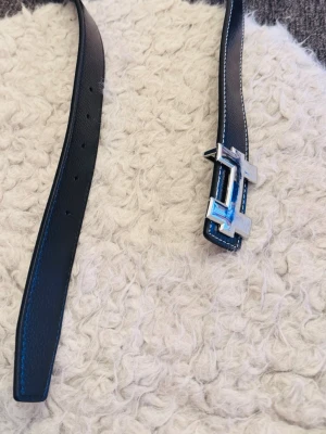 Svart skärp med silverfärgat H-spänne - Snyggt svart bälte i skinn med ett stort, silverfärgat H-format spänne. Bältet har vita kontrastsömmar längs kanterna och flera hål för justerbar passform. Perfekt accessoar för att lyfta vilken outfit som helst.