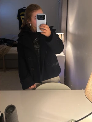 Svart skepparkavaj med dubbelknäppning - Stilren svart skepparkavaj med klassisk dubbelknäppning och bred krage. Jackan har dekorativa knappar vid ärmsluten och är tillverkad i ett tjockare tyg som känns ulligt. Perfekt för dig som vill ha en tidlös och snygg look.