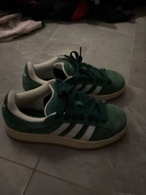 Gröna Adidas sneakers i mocka - Snygga gröna Adidas sneakers i mocka med klassiska vita ränder på sidorna och vit sula. Skorna har lågt skaft och gröna skosnören. Perfekta för dig som gillar retrostil och vill sticka ut lite extra.