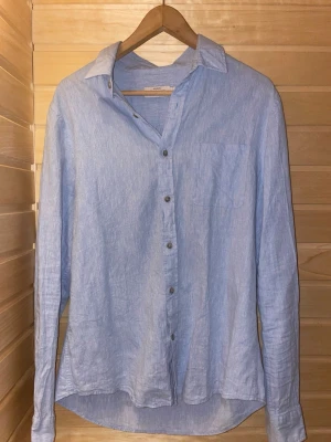 Ljusblå linneskjorta - Snygg ljusblå skjorta i linne med klassisk krage och knäppning framtill. Skjortan har lång ärm och en bröstficka, perfekt för en avslappnad men stilren look.