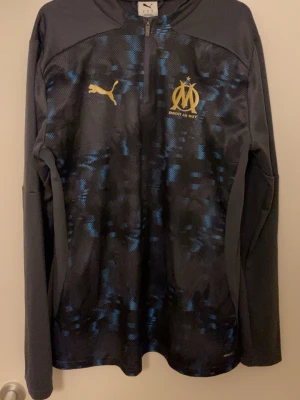 Marseille tröja  - Säljer en svart långärmad träningströja från Puma med Olympique de Marseille-logga i guld på bröstet. Tröjan har blått mönster framtill och baktill, halv dragkedja och sportig passform. Perfekt för fotbollsträning eller gymmet.