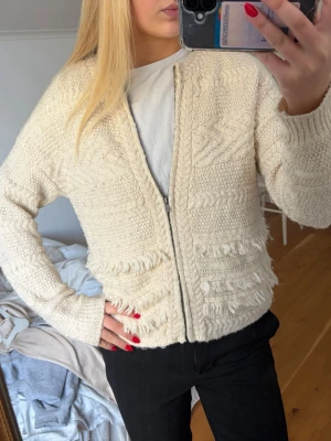 Krämvit stickad kofta med dragkedja - Snygg krämvit stickad kofta med dragkedja framtill. Koftan har ett unikt mönster med struktur och fransdetaljer, samt långa ärmar och ribbade muddar. Perfekt för lager-på-lager och en chill look.