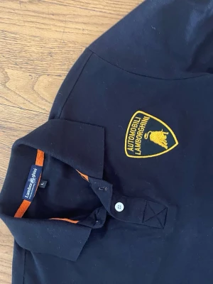 Svart Lamborghini pikétröja - Snygg svart pikétröja från Lamborghini med gul broderad logga på bröstet och orange detaljer i kragen. Klassisk pikéknäppning med två knappar och korta ärmar. Perfekt för dig som gillar sportiga och exklusiva plagg.