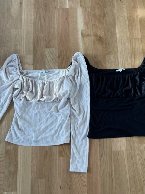 Två ribbade toppar med volang - Säljer två snygga ribbade toppar med volangdetalj framtill. Ena toppen är beige och den andra svart, ena har lång ärm och och den andra har kort ärm, fyrkantig urringning. Perfekta att styla med jeans eller kjol för en trendig look.