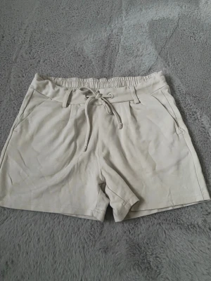 Mjuka beiga shorts. - Snygga beige shorts med elastisk midja och snörning framtill. Shortsen har fickor på sidorna och dekorativa bakfickor. Perfekta för varma dagar och enkel att matcha med olika toppar.