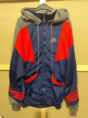 Adidas hoodie i blått, rött och grått - Snygg hoodie från Adidas i marinblått med röda detaljer och grå huva. Dragkedja framtill, två fickor och broderad Adidas-logga på bröstet. Muddar vid ärmslut och nederkant. Perfekt för en sportig och avslappnad stil.