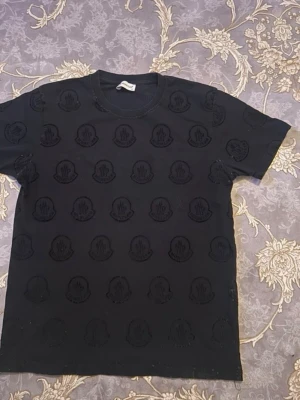 Svart Moncler t-shirt med logomönster - Snygg svart t-shirt från Moncler med upprepade ton-i-ton loggor över hela plagget. Klassisk rund halsringning och korta ärmar. Tillverkad i mjuk bomull som känns skön mot huden. Perfekt för dig som gillar stilrena plagg med exklusiv känsla.