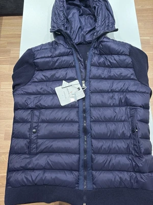 Mörkblå dunväst från Moncler - Snygg mörkblå dunväst från Moncler med huva och dragkedja framtill. Västen har två fickor med knappstängning och ribbade detaljer vid ärmöppning och nederkant. Perfekt för lager-på-lager och streetstyle. Materialet är glansigt och quiltat för en modern look.