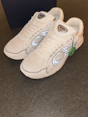 Vita Dior B30 sneakers - Säljer ett par vita Dior B30 sneakers med reflekterande silverdetaljer och meshpaneler. Skorna har en chunky sula och tydlig Dior-logga på sidan. Klassisk low-top modell med vita skosnören och autentiseringsbricka från StockX.
