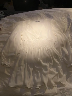 Vit klänning - Snygg vit omlottklänning i luftig chiffong med smockad midja. Klänningen har långa ärmar med resår vid ärmslut och en feminin v-ringning. Jag som är 177cm lång så går den ner till ca mitten av låret.