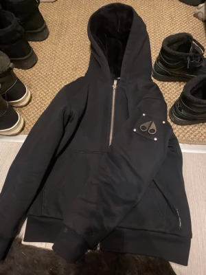  Moose Knuckles Bunny svart päls - Säljer en svart hoodie från Moose Knuckles med dragkedja och stor huva. Hoodien har en snygg metallogo på ärmen och rymliga fickor framtill. Tillverkad i mjukt och tjockt material som känns riktigt lyxigt. Perfekt för dig som gillar streetwear och vill ha något stilrent.