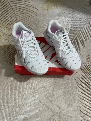 Nike Air Max Plus vita sneakers - Nike Air Max Plus sneakers i vitt med rosa och silverdetaljer. Ovandel i mesh och syntet, synlig Air Max-dämpning i sulan och klassisk Nike-logga på sidan. Snygg design med coola detaljer och grå yttersula. Perfekta för dig som gillar streetwear och sportig stil.