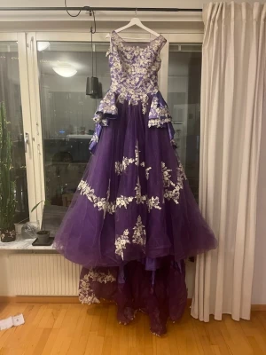 Lila långklänning med spetsdetaljer - Unik lila långklänning med voluminös kjol och vackra vita spetsapplikationer. Klänningen har ärmlös design, markerad midja och flera lager tyg som ger extra volym. Perfekt för bal eller festliga tillfällen. Ingen synlig logga på märket. Aldrig använd