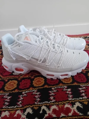 Nike Air Max Plus vita sneakers - Nike Air Max Plus sneakers i helvitt med mesh- och syntetmaterial, ikoniska vågiga detaljer och synliga Air-enheter med orange inslag i sulan. Skorna har en chunky sula och klassisk snörning. Perfekta för dig som gillar streetwear och sportig stil.
