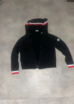Svart Moncler hoodiejacka med huva - Svart hoodiejacka från Moncler med dragkedja och huva. Jackan har Moncler-logga på ärmen och ribbade muddar med röd, vit och blå rand. Tillverkad i mjuk bomull och har en clean, sportig look. Perfekt för dig som gillar streetwear och vill ha något stilrent.