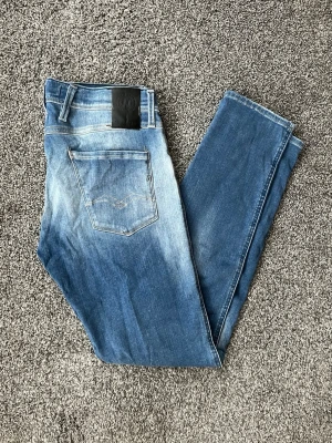 Blå slim fit jeans från Replay - Säljer ett par blå slim fit jeans från Replay med snygga slitningar och tvättade detaljer framtill. Klassisk femficksmodell med läderpatch bak i midjan. Jeansen har normal midja och smala ben. Perfekta för en trendig och avslappnad stil.