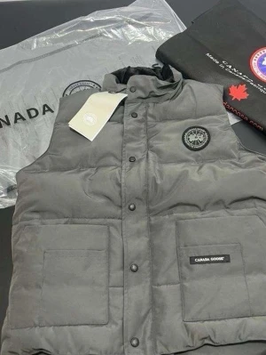 Grå dunväst från Canada Goose - Snygg grå dunväst från Canada Goose med hög krage, två stora framfickor och tryckknappar. Västen har en diskret logotyp på bröstet och en patch med märkesnamnet på fickan. Perfekt för lager-på-lager och streetstyle.