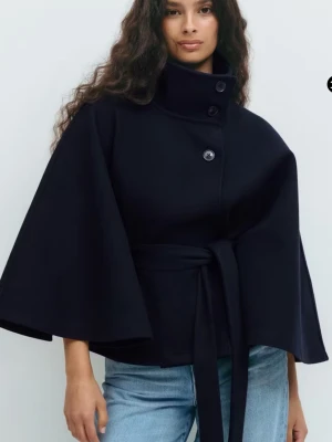 Marinblå cape-kappa med knytband - Stilren marinblå cape-kappa med hög krage, knappar framtill och knytband i midjan. Kappan har vida ärmar och en kortare, boxig passform. Perfekt för dig som vill ha en trendig och elegant look. Materialet ser ut att vara ullblandning. Sitter perfekt på mig som är en smål 🤍