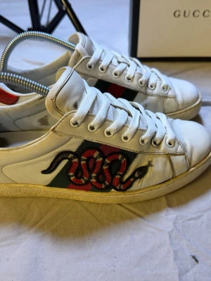 Gucci Ace sneakers – Klassiska ormen – Box & dustbags – Äkthetsbevis - Säljer ett par Gucci Ace sneakers med den klassiska ormen, ett av Guccis mest ikoniska motiv. Skorna kommer med box, dustbags samt äkthetsbevis från Mytheresa, vilket gör köpet extremt tryggt.  En tidlös designer-sneaker som aldrig går ur stil och passar till allt från jeans till kostymbyxor.  • Märke: Gucci • Modell: Ace • Detalj: Klassiska ormen • Tillbehör: Box, dustbags, äkthetsbevis