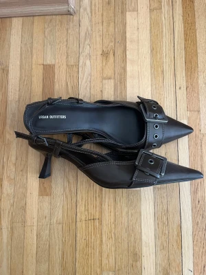Bruna kitten heels med spänne Urban Outfitters - Snygga bruna kitten heels från Urban Outfitters med spetsig tå, breda remmar och stora silverspännen framtill. Skorna har öppen häl och justerbara remmar. Tillverkade i konstläder med markerade sömmar för en cool look. Aldrig använda