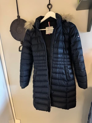 Marinblå lång dunjacka Tommy Hilfiger - Snygg marinblå lång dunjacka från Tommy Hilfiger med huva och fuskpälsdetalj. Jackan har quiltad design, dragkedja framtill och diskret logga på ärmen. Perfekt för kalla vinterdagar och riktigt stilren look.