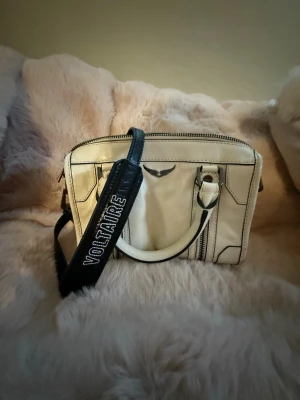 Vit handväska från Zadig & Voltaire - Snygg vit handväska i läder från Zadig & Voltaire med svarta detaljer och bred axelrem med logga. Väskan har dragkedja upptill, innerfack och stilrena metalldetaljer. Perfekt för dig som vill ha en trendig och praktisk accessoar.