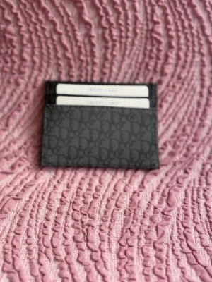 Svart  korthållare i läder - Snygg svart korthållare med diskret mönster av märkets logga över hela ytan. Tillverkad i läder och har flera fack för kort. Perfekt för dig som vill ha en stilren och lyxig accessoar i fickan.