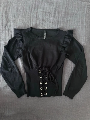 Svart topp med snörning och volang - Snygg fin stickad svart tröja med långa ärmar och cool snörning framtill. Perfekt för en trendig look. Passar bra till nästan allt och sitter jätte skönt, funkar som både S och M.