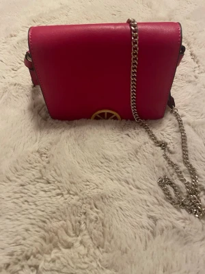 Rosa axelremsväska med citrondetalj - Snygg rosa handväska med guldkedja och dekorativa rosetter på sidorna. Väskan har en unik citronskiva i metall på framsidan som detalj. Perfekt för dig som vill sticka ut med en färgglad accessoar. Rektangulär form och stilren design.