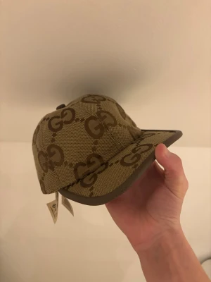 Gucci beige keps med GG-mönster - Snygg beige keps från Gucci med det klassiska GG-monogrammet i brunt över hela kepsen. Kepsen har böjd skärm, justerbart spänne bak och är tillverkad i canvasmaterial med läderdetaljer. Perfekt för dig som gillar exklusiva accessoarer. Använda inget mycket vinter tider va ska man säga liksom och om priset är för högt kan det diskuteras för de ska ligga bra för oss båda 
