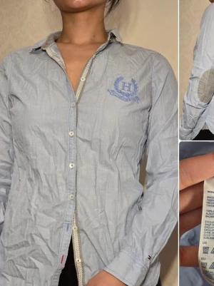 Blå skjorta från Tommy Hilfiger - Klassisk ljusblå skjorta från Tommy Hilfiger med broderad logga på bröstet och kontrasttyg på insidan av kragen. Skjortan har knappar framtill, lång ärm med förstärkta armbågslappar i grått och är tillverkad i 100% bomull. Finns en liten fläck vid kragen