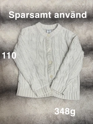 Vit kabelstickad kofta Polarn o pyret barn - Mysig vit kabelstickad kofta från Polarn o pyret med rund halsringning och stora knappar framtill. Koftan har ribbade muddar vid ärmslut och nederkant, perfekt för lager på lager och en klassisk look.🌸 Sparsamt använd. Storlek 110