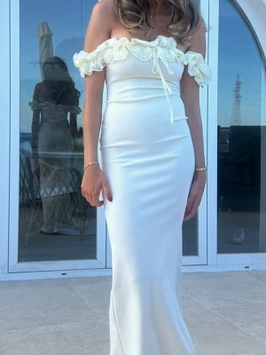 Vit balklänning med volang - Elegant vit långklänning med offshoulder och volangdetaljer upptill. Klänningen har en smickrande slim passform och knyt framtill vid bysten. Perfekt för sommarens speciella tillfällen.