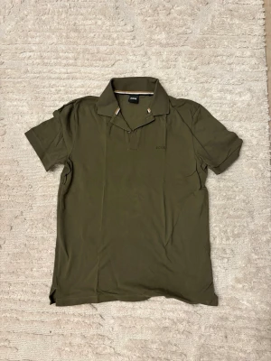 Olivgrön piké BOSS - Snygg olivgrön pikétröja från BOSS med klassisk krage och korta ärmar. Tröjan har diskret BOSS-logga broderad på bröstet och kontrastdetaljer i kragen. Perfekt för en stilren och avslappnad look.
