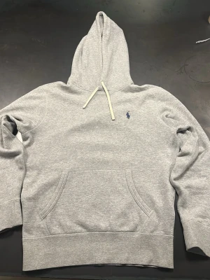 Grå hoodie från Polo Ralph Lauren - Klassisk grå hoodie från Polo Ralph Lauren med broderad marinblå logga på bröstet. Hoodien har huva med vita dragsnören och en stor magficka framtill. Tillverkad i mjuk bomullsblandning och har ribbade muddar vid ärmslut och nederkant. Hoodie:n är i mycket bra skick och är väl omhändertagen. Bara att skriv vid funderingar