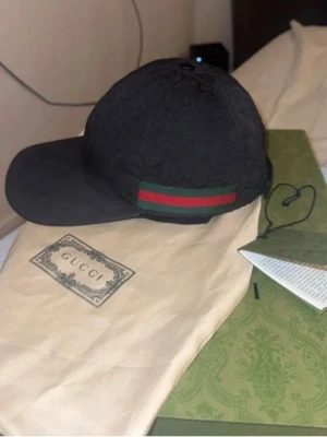 Svart keps från Gucci med GG-mönster - Snygg svart keps från Gucci med klassiskt GG-monogram i tyget och en grön-röd rand på sidan. Kepsen har böjd skärm och är tillverkad i ett tygmaterial med exklusiv känsla. Perfekt accessoar för att lyfta din streetwear-look.