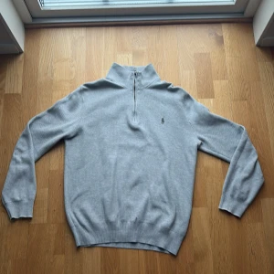 Ljusgrå stickad tröja från Ralph Lauren - Ljusgrå stickad tröja från Ralph Lauren med halv dragkedja och hög krage. Tröjan har långa ärmar, ribbstickade muddar och en liten broderad logotyp på bröstet. Perfekt för lager-på-lager och stilrena outfits.