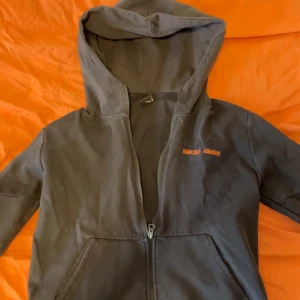 Svart hoodie från Emporio Armani Ea7 - Svart hoodie med dragkedja från Emporio Armani. Märkets logga i orange på bröstet och en reflexdetalj på ärmen. Klassisk huva och två fickor framtill. Perfekt för en avslappnad och stilren look.