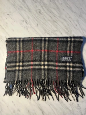 Burberry halsduk 100% kashmir  - Säljer en klassisk rutig halsduk från Burberrys of London i 100% mjuk kashmir. Den har en snygg grå bas med vita och röda rutor samt traditionella tvinnade fransar i ändarna. Halsduken är märkt med Burberrys of London