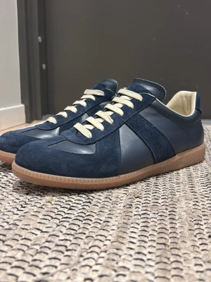 Maison Margiela gats  - KOM MED PRISFÖRSLAG. Snygga marinblå sneakers med ovandel i mocka och skinn, beige snörning och klassisk gummisula. Skorna har en stilren design med rund tå och kontrasterande detaljer. Perfekta för dig som gillar en clean och tidlös look.
